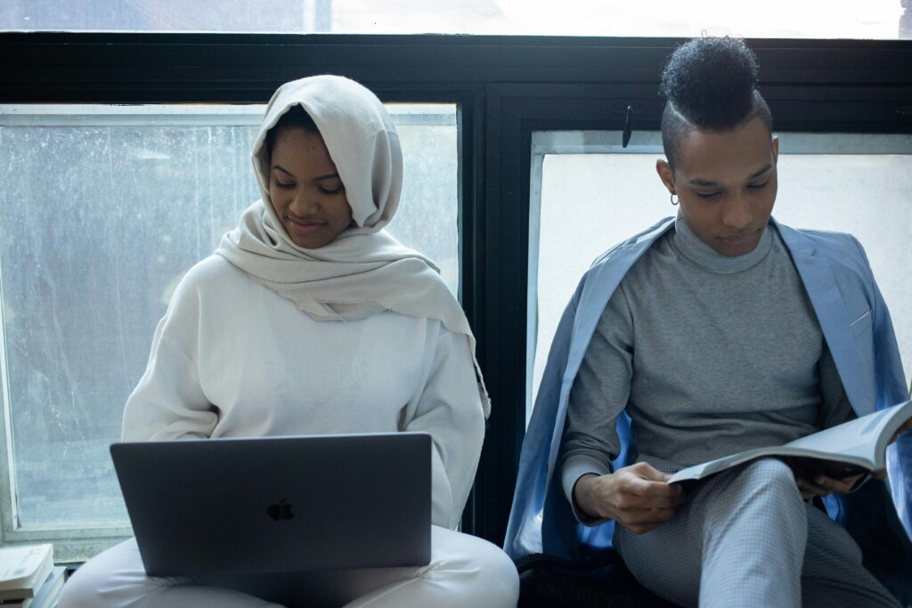 Concentrated African American woman in hijab working on laptop near classmate reading textbook during exam preparation